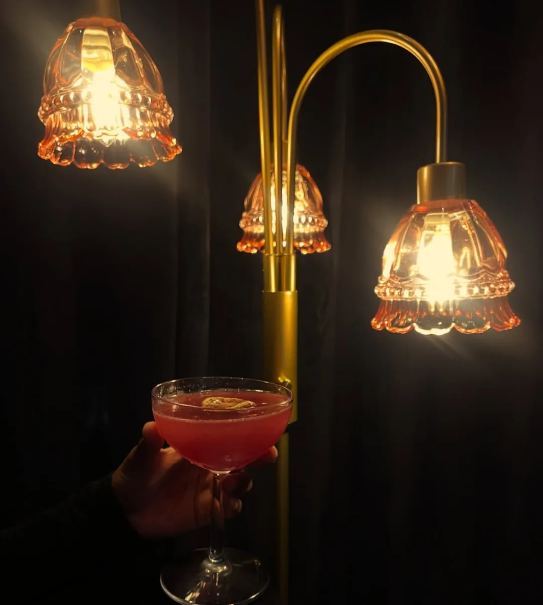 A blush-pink cocktail held up beneath glowing amber glass lamps in the lounge at Lee's Coffee in Birmingham