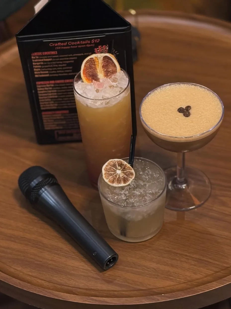 Three handcrafted cocktails and a microphone on a wooden table beside the Lee's cocktail menu